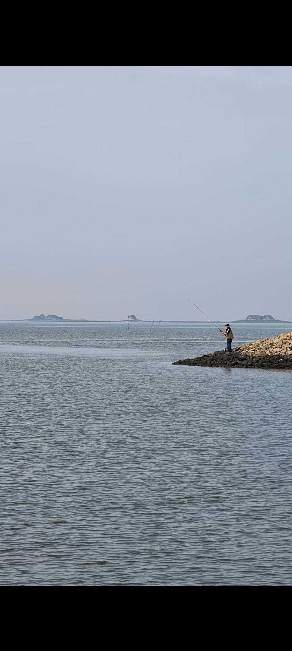 Ein Angler steht auf einem Steinwall an der Nordsee bei Hochwasser, im Hintergrund ist die Hallig Nordstrandischmoor am Horizont zu sehen. 