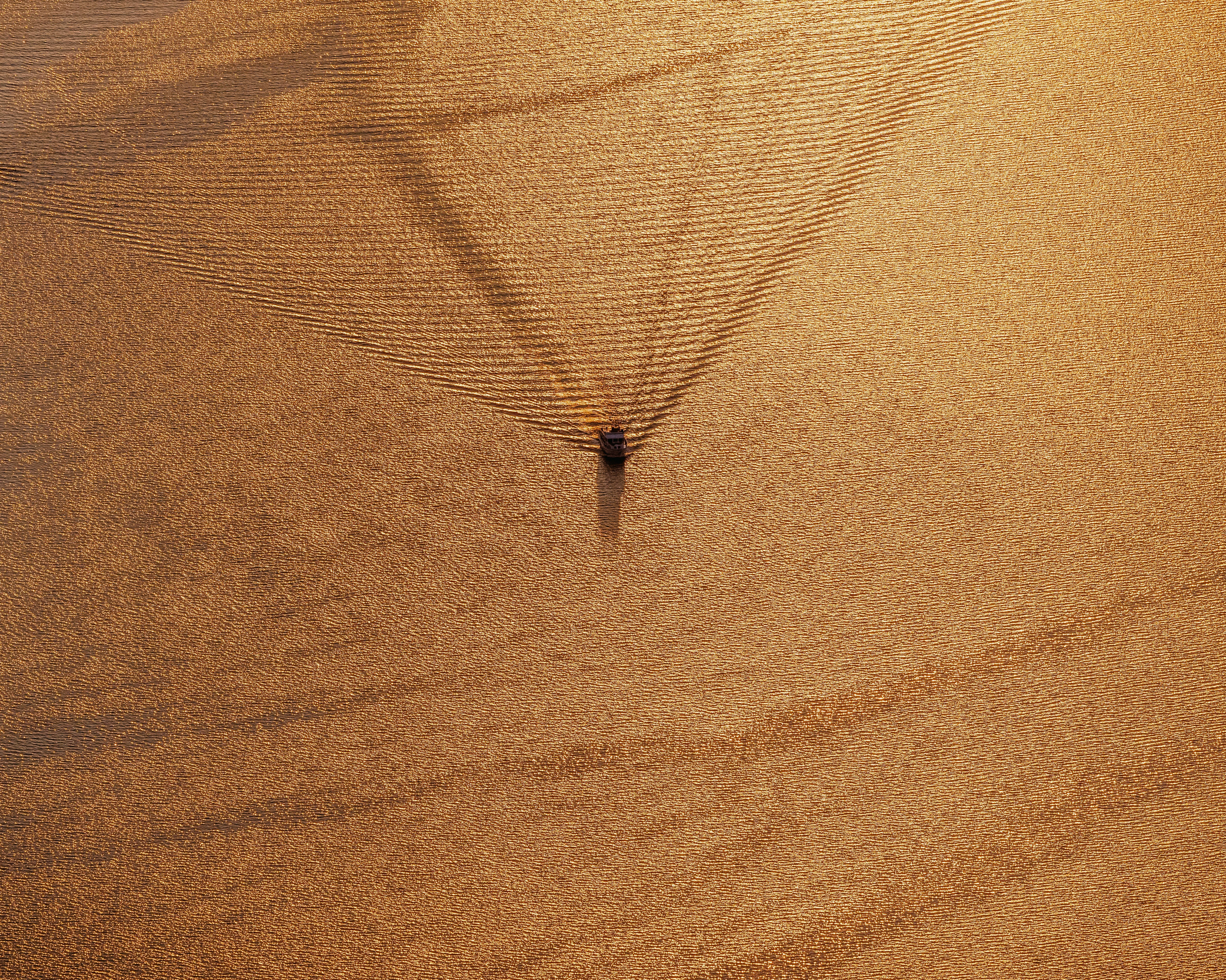 Vogelperspektive auf einen Fischkutter, der im Sonnenuntergang fährt, mit goldenen Lichtreflexen auf dem Wasser.