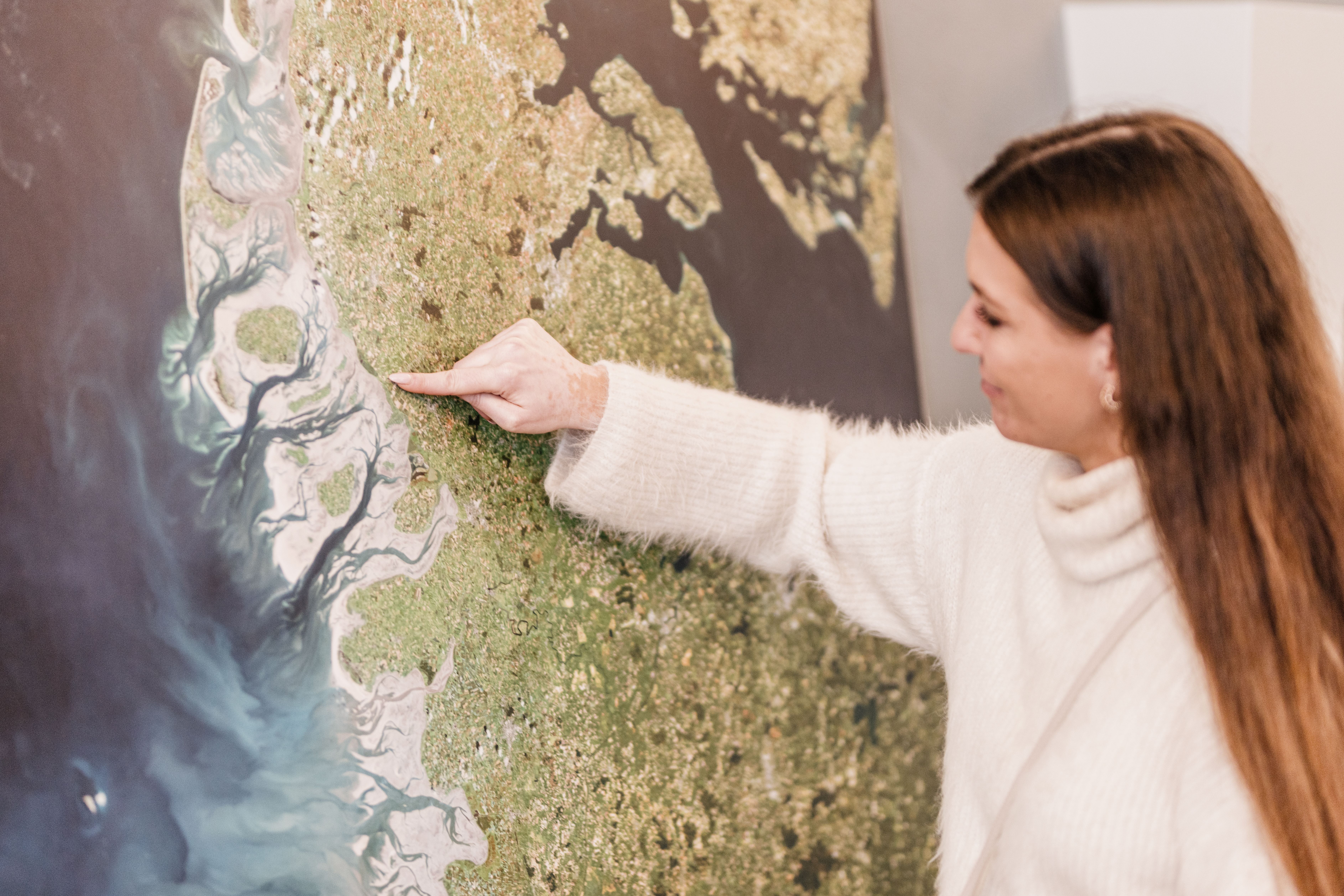 Eine Frau zeigt mit ihrem Zeigefinger auf einen Punkt auf einer Karte der Nordseeküste Schleswig-Holsteins, die an einer Wand hängt. Verlinkung zur Unterseite "Museen und Kultur".
