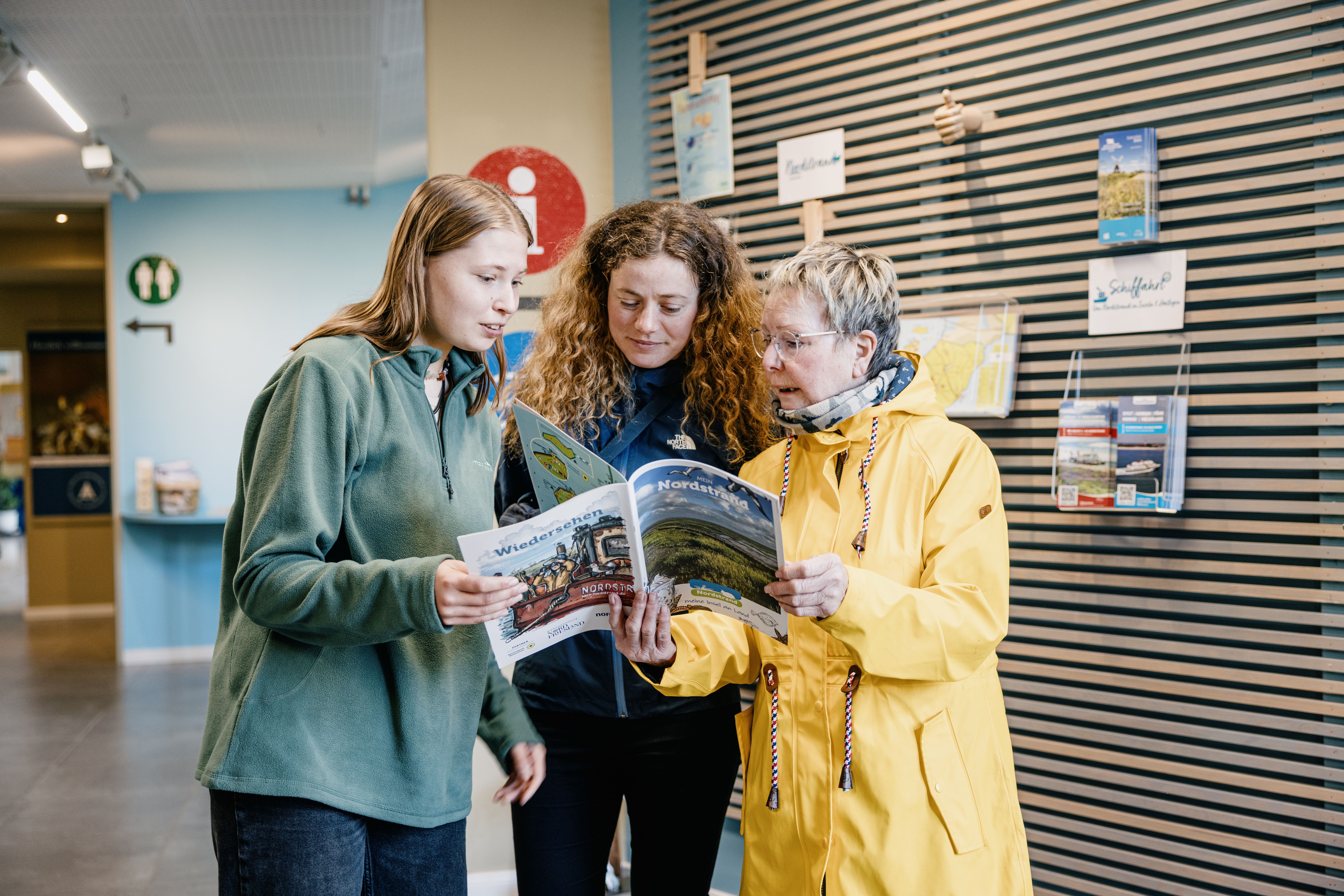 Drei Frauen in der Tourist-Information Nordstrand und schauen in das Urlaubsmagazin von Nordstrand. Verlinkung auf ein neues Fenster mit der PDF-Datei des Urlaubsmagazins.