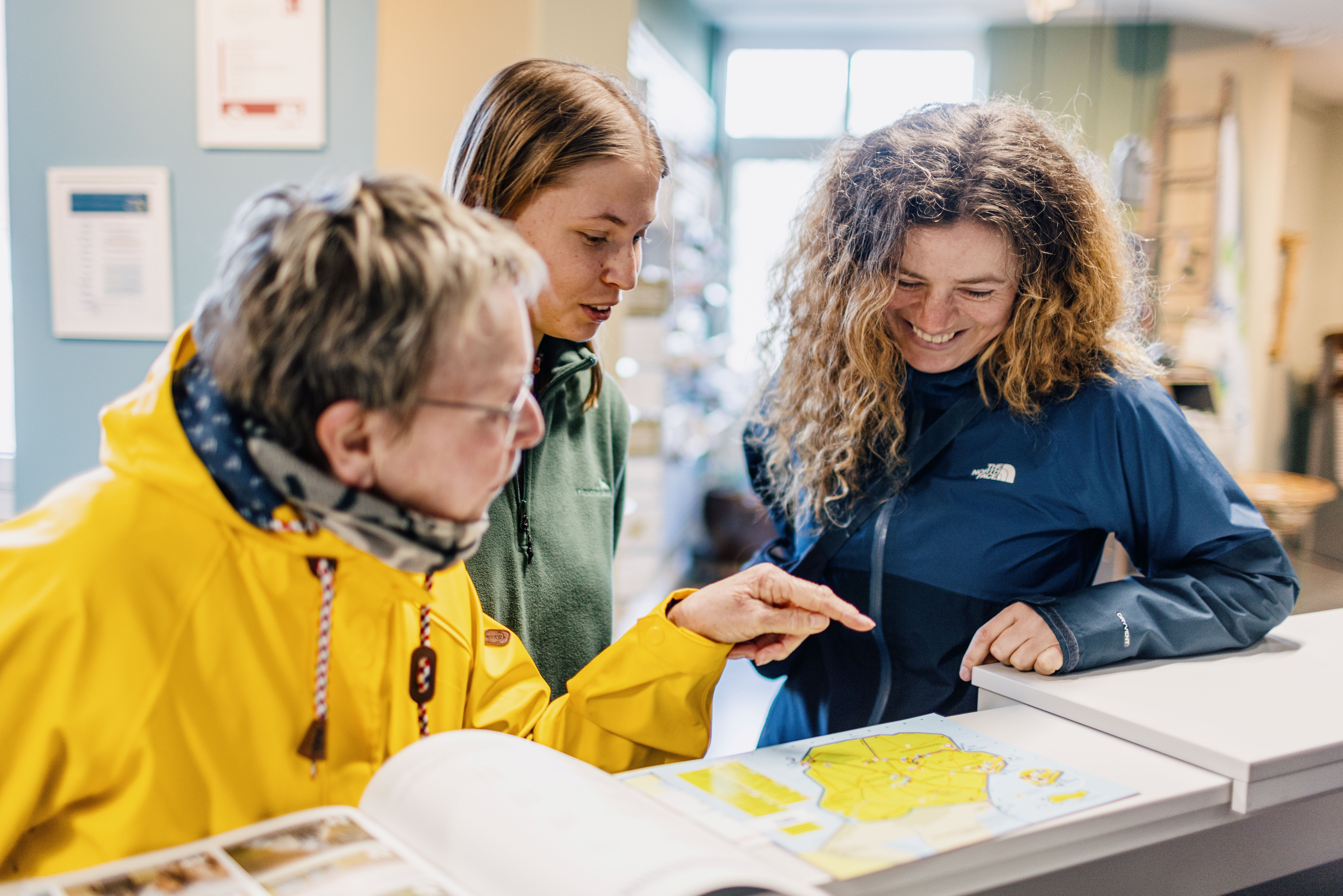 Drei Frauen am Schalter der Tourist-Informationen auf Nordstrand, Verlinkung auf Unterseite "Magazine & Broschüren".
