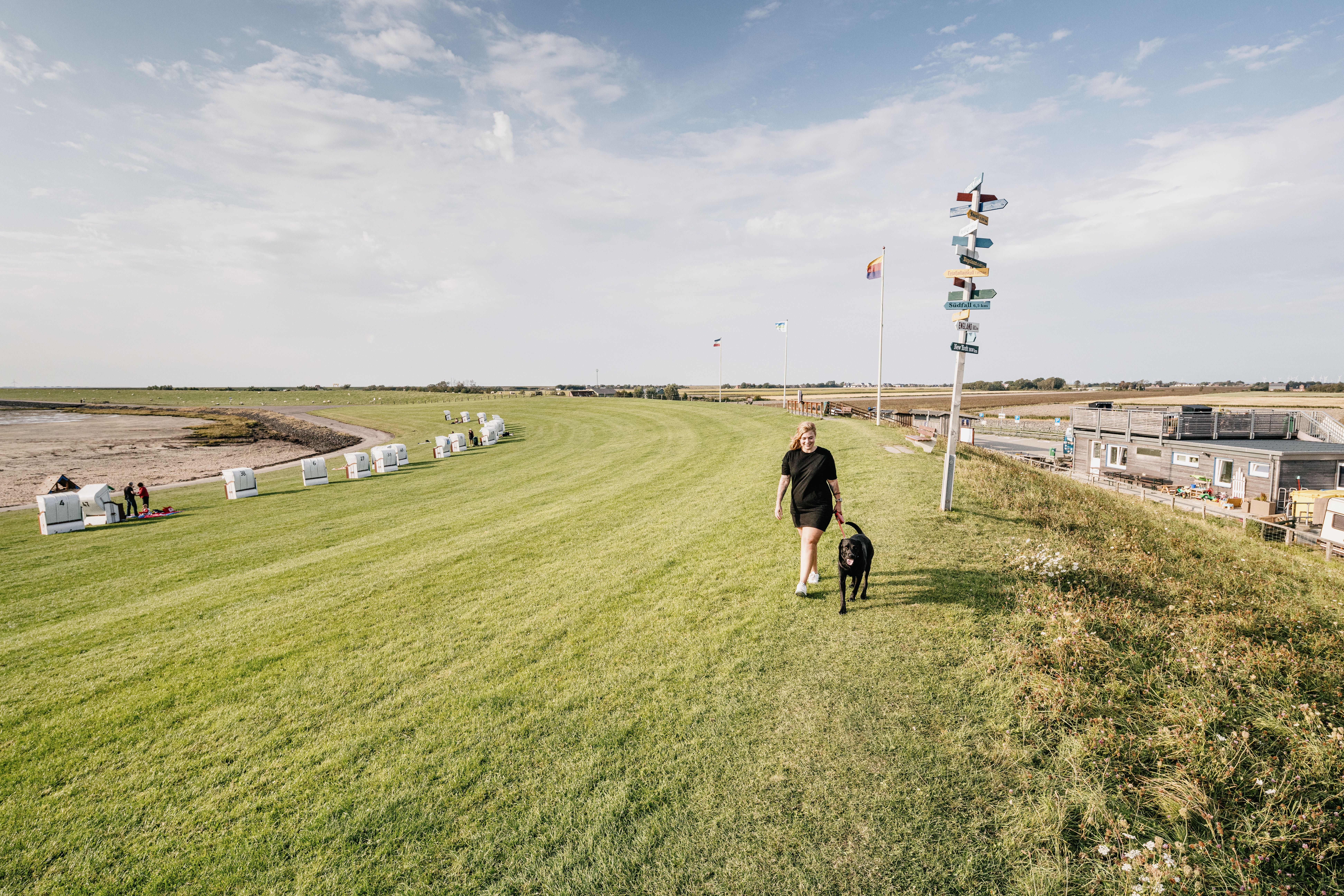 Eine Frau in schwarzer Kleidung läuft auf einem Deich mit ihrem angeleinten, schwarzen Hund. 