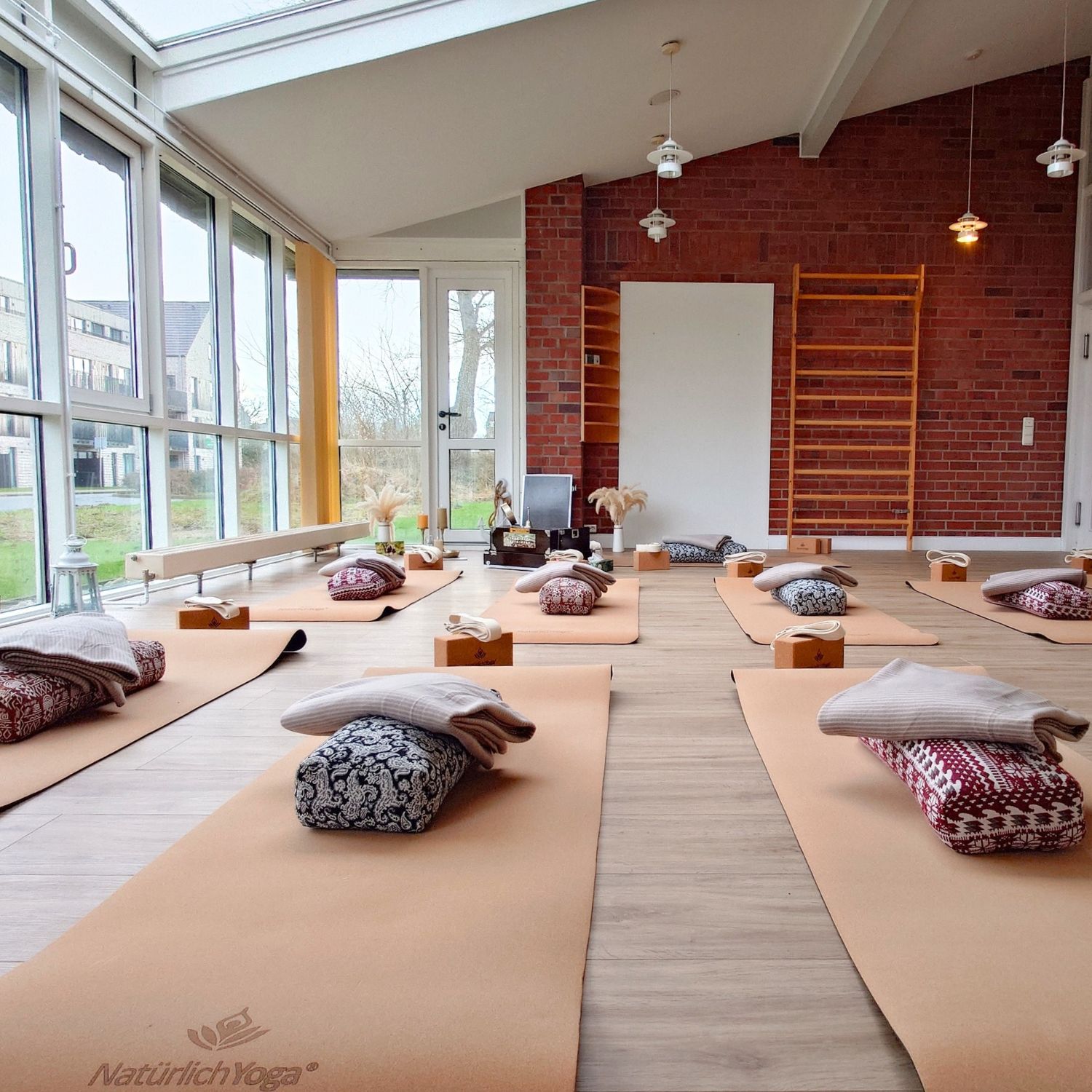 In einem Raum liegen 6 Yogamatten mit Kissen und Decken, an der linken Seite ist eine Glasfront.
