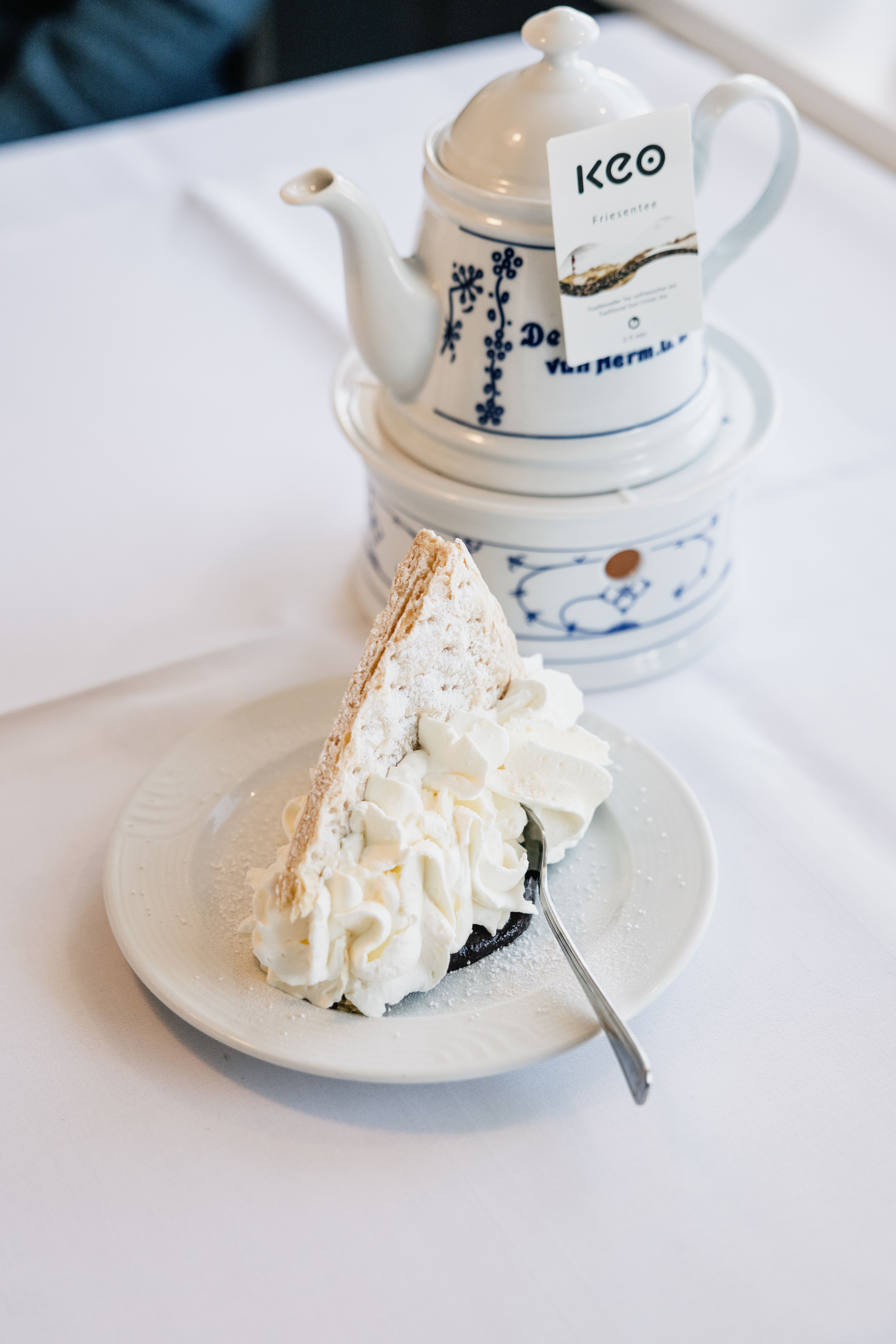Eine Teekanne steht neben einem Teller mit einem Stück Friesentorte auf einer weißen Tischdecke. 