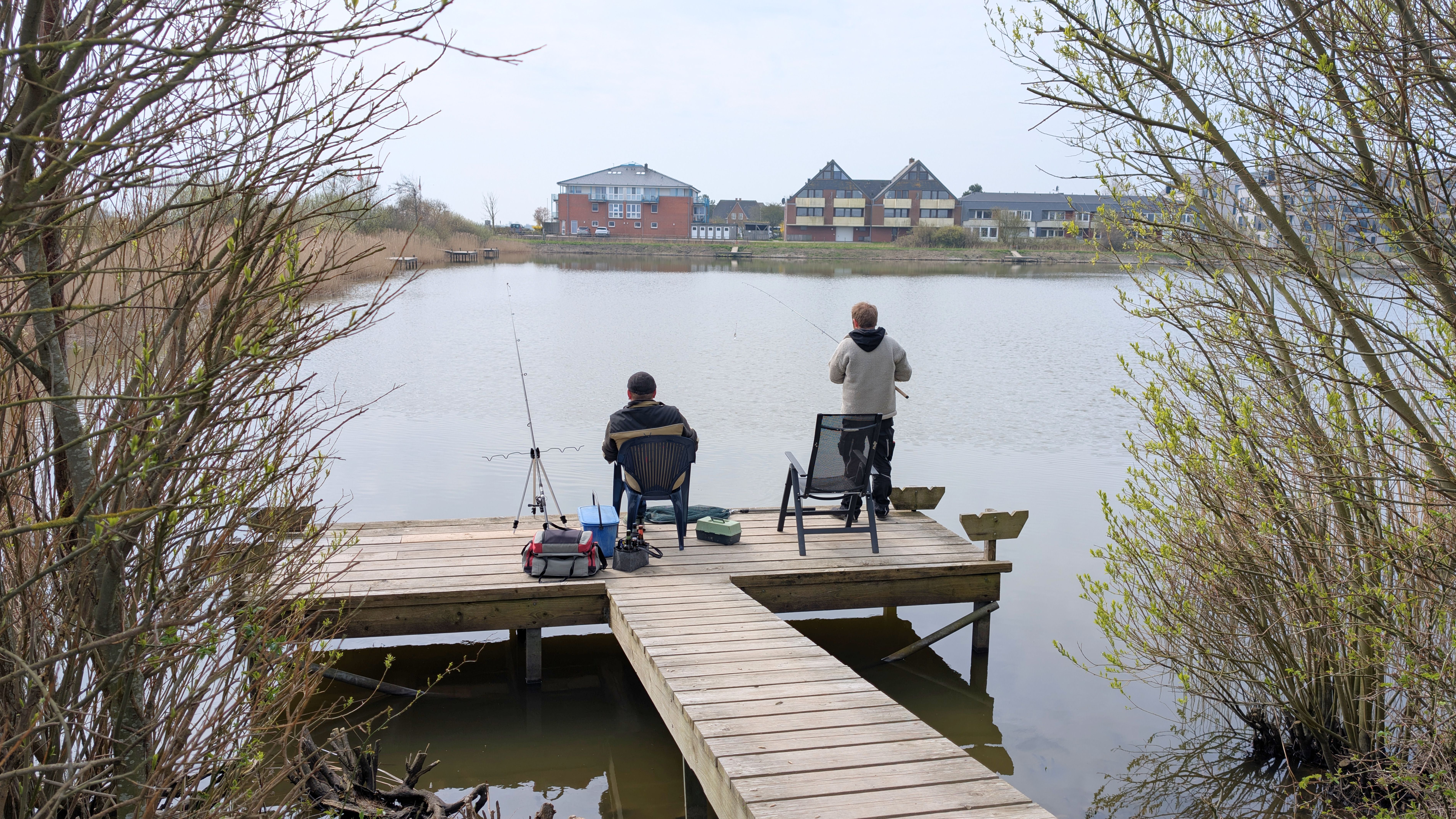 Zwei Angler sitzen auf einem Steg an einem See. 