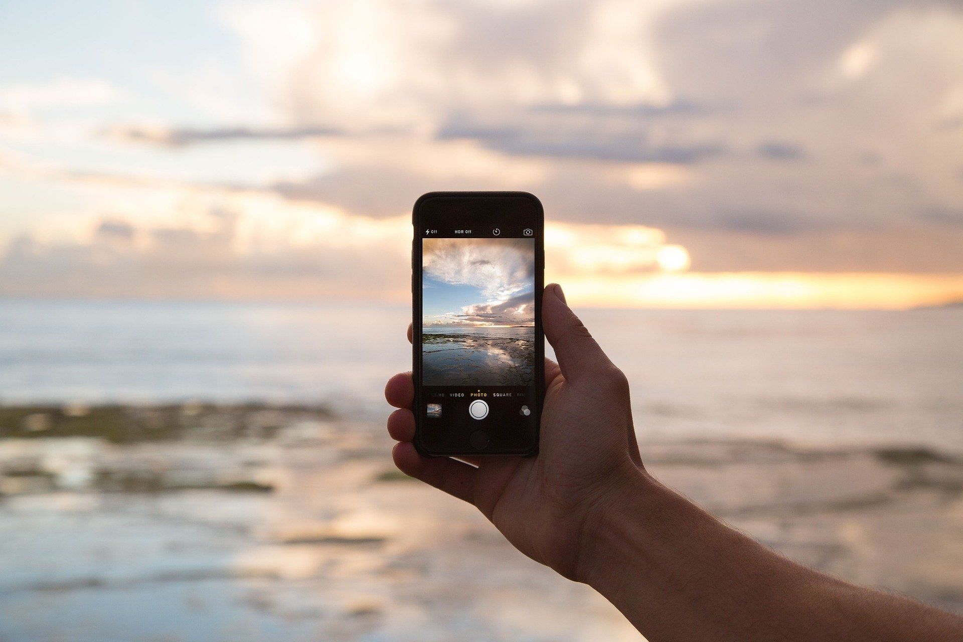 Eine Hand hält ein Smartphone im Kameramodus auf die Nordsee gerichtet bei Sonnenuntergang. 