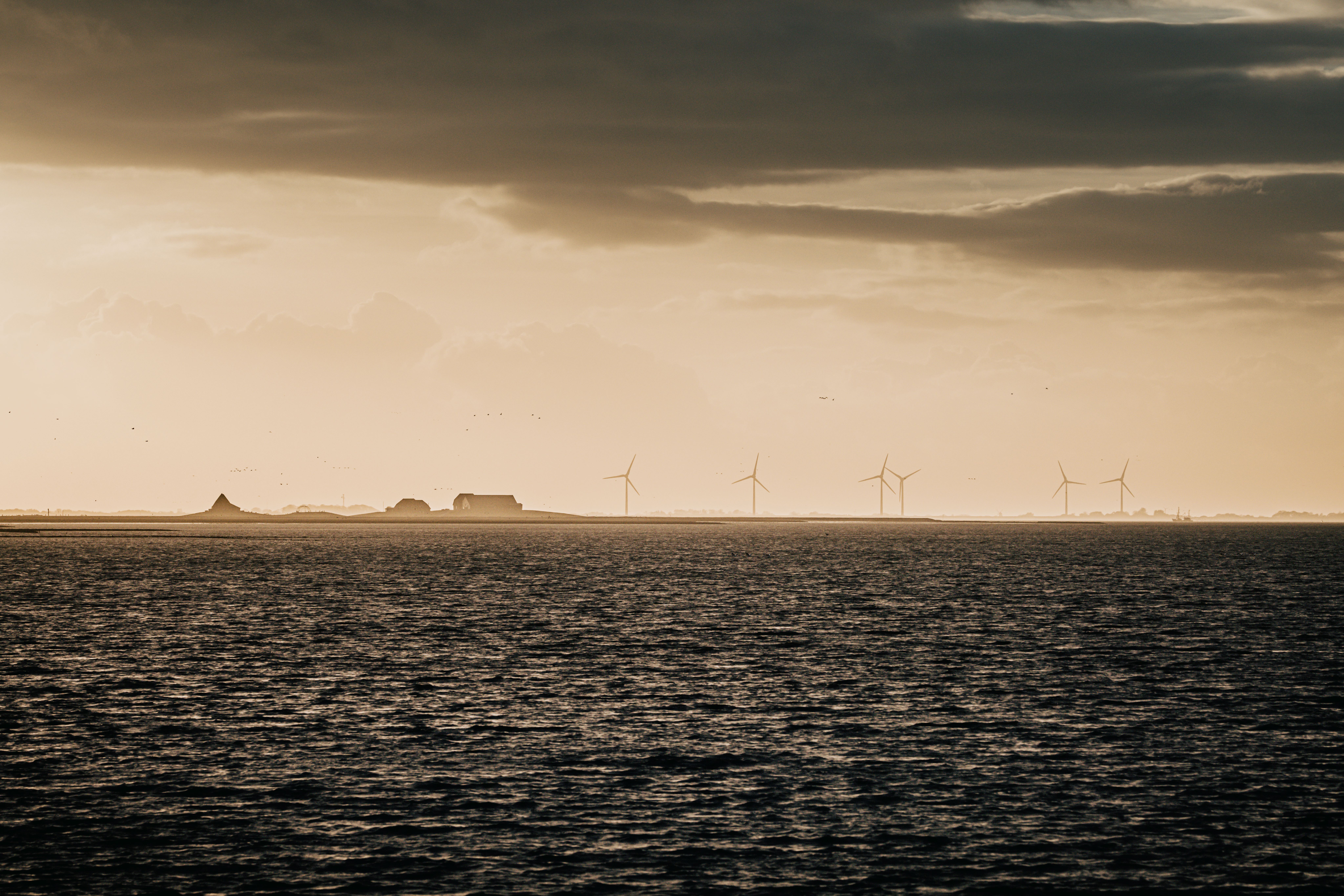 Die Nordsee bei Hochwasser und beim Sonnenuntergang, am Horizont die Umrisse von Hallig Nordstrandischmoor und Windkraftanlagen. Verlinkung auf "Unterkünfte am Meer"