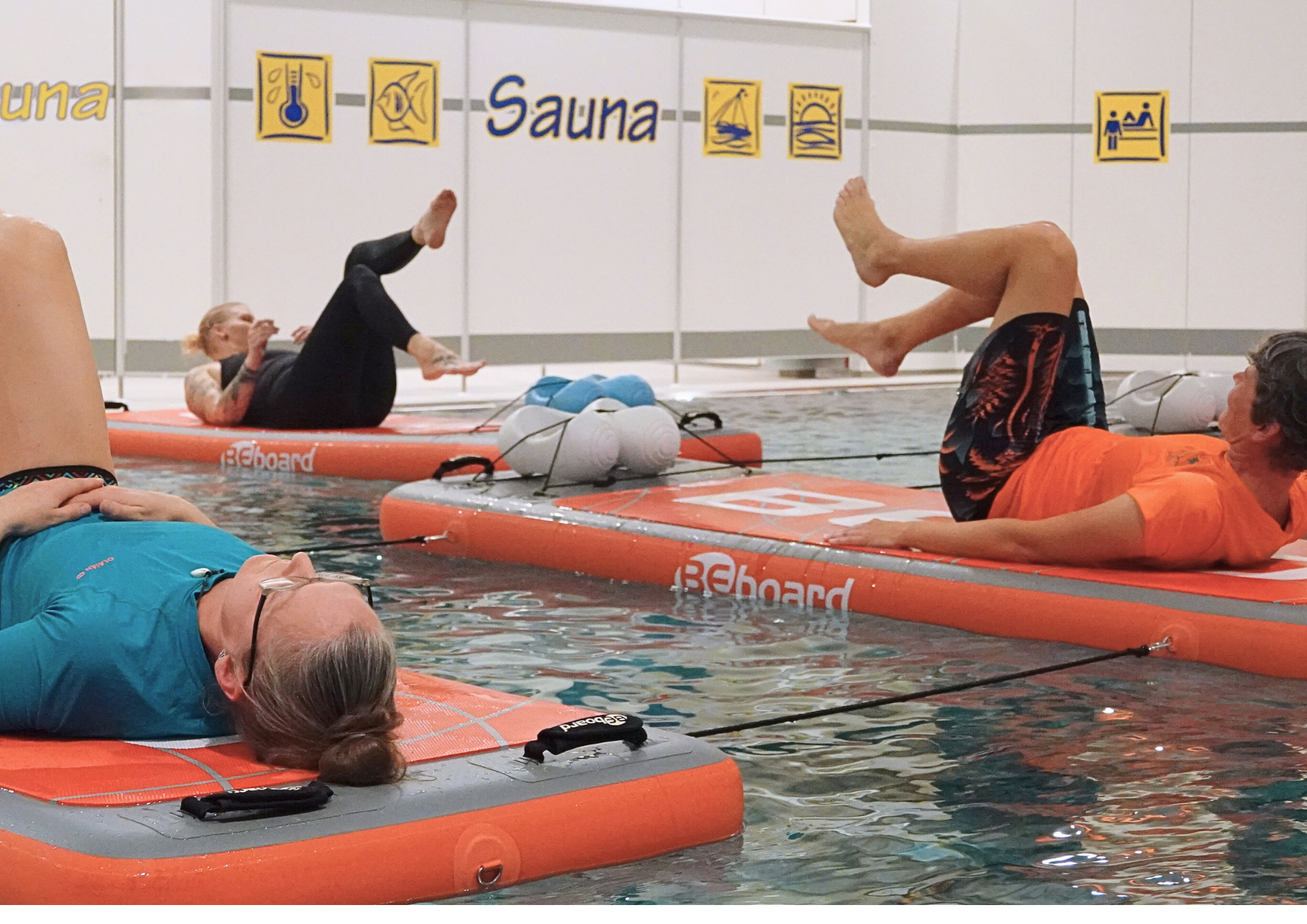 Personen machen Sportübungen auf einem aufblasbaren Brett in einem Schwimmbecken. 