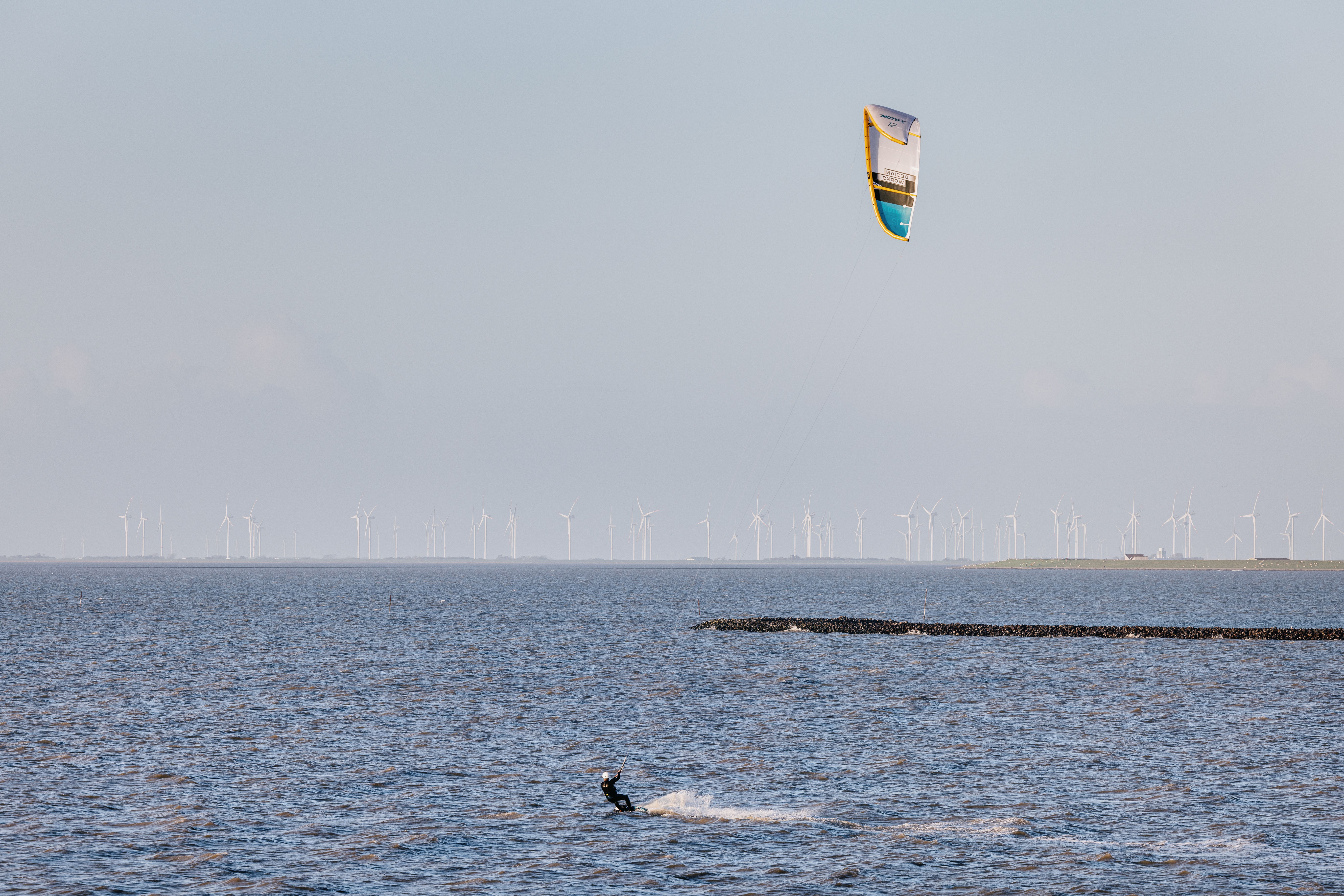Ein Kiter fährt auf dem Meer, im Hintergrund Windkraftanlagen. 