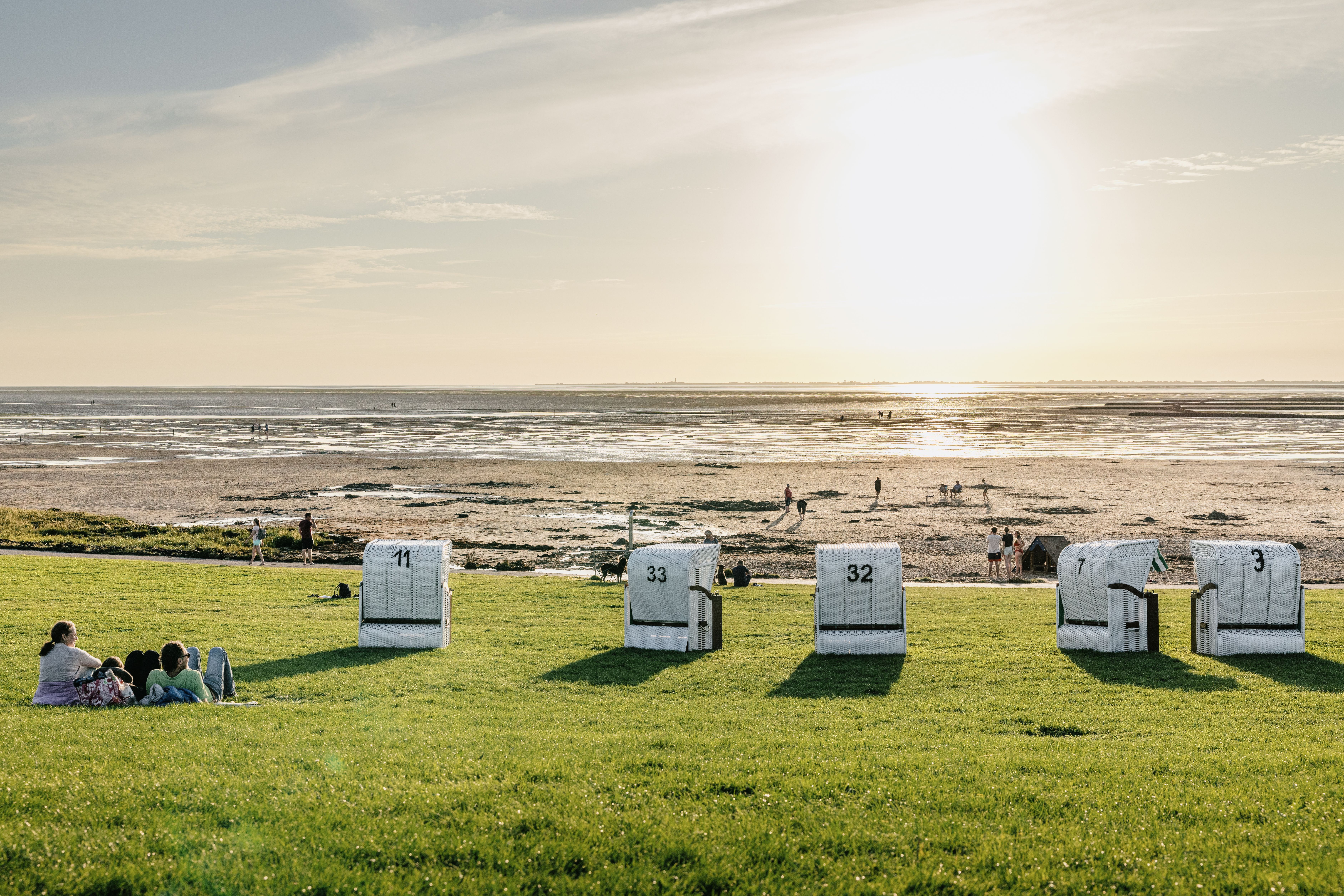 Ein Pärchen liegt neben 5 weißen Strandkörben am Gründeich bei Niedrigwasser und Sonnenuntergang. 