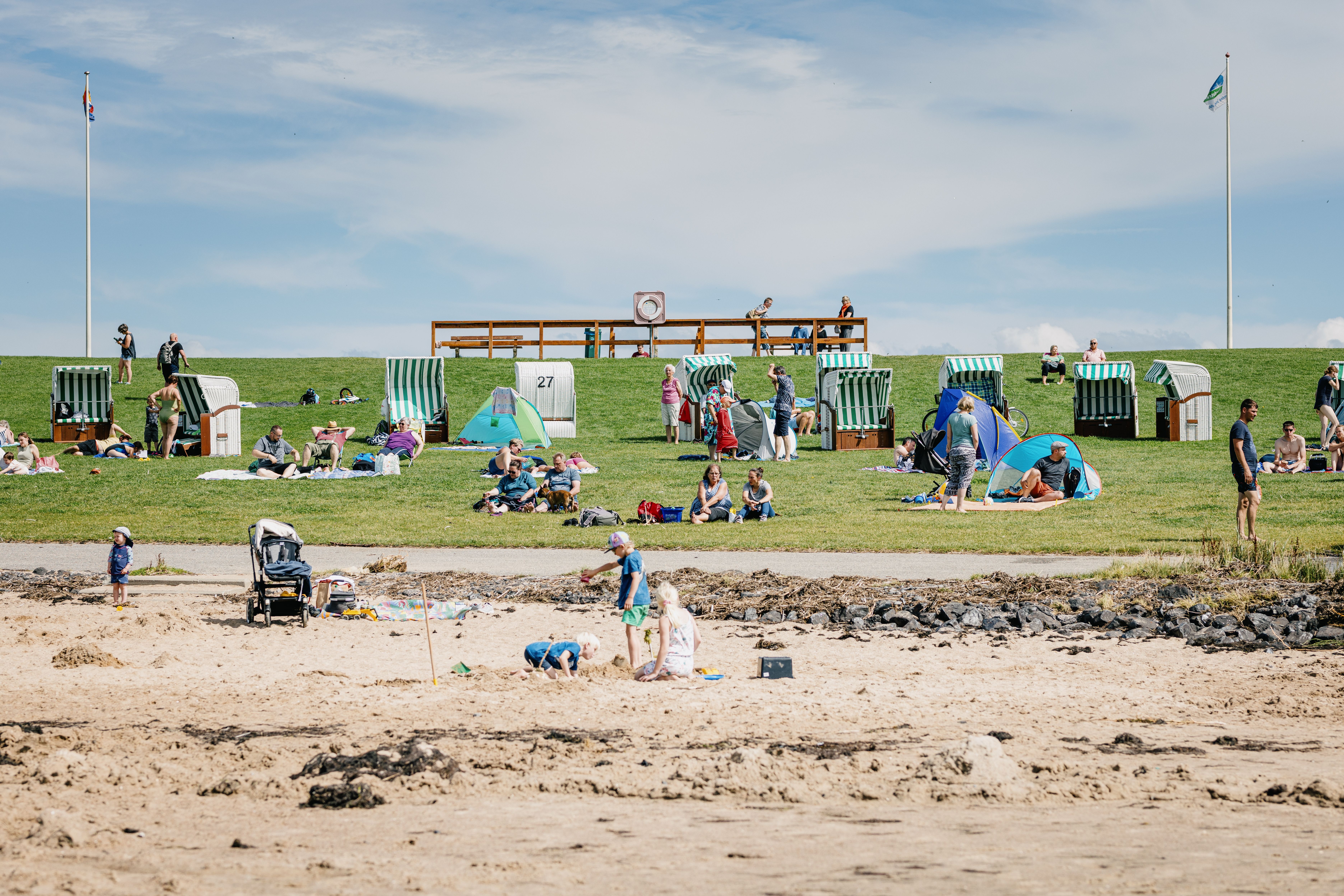 Kinder spielen im Sand, auf dem Deich sitzen Personen im Strandkorb oder im Gras auf dem Deich. 