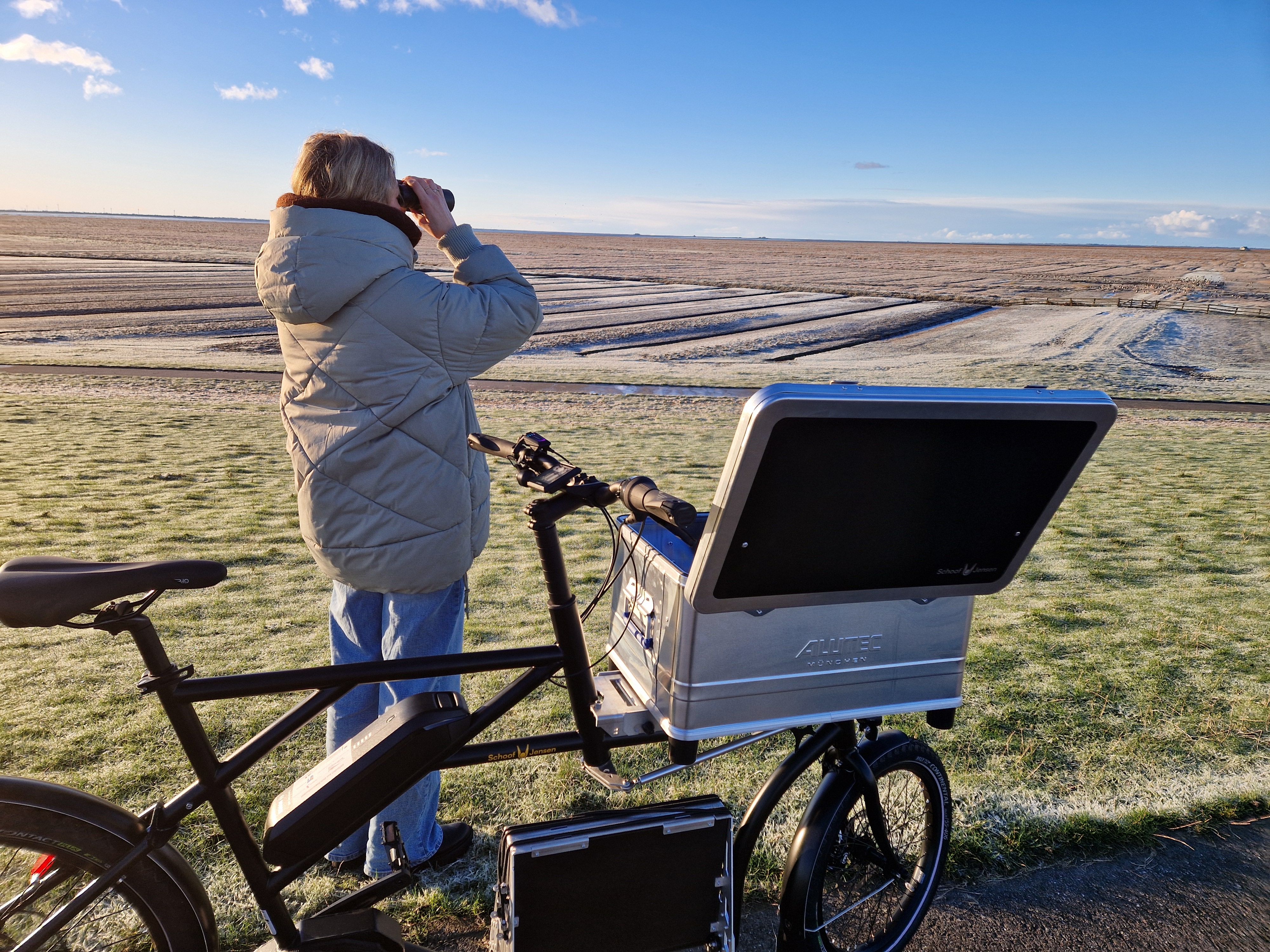 Eine Frau steht an Salzwiesen mit einem Lastenrad und schaut durch ein Fernglas. 