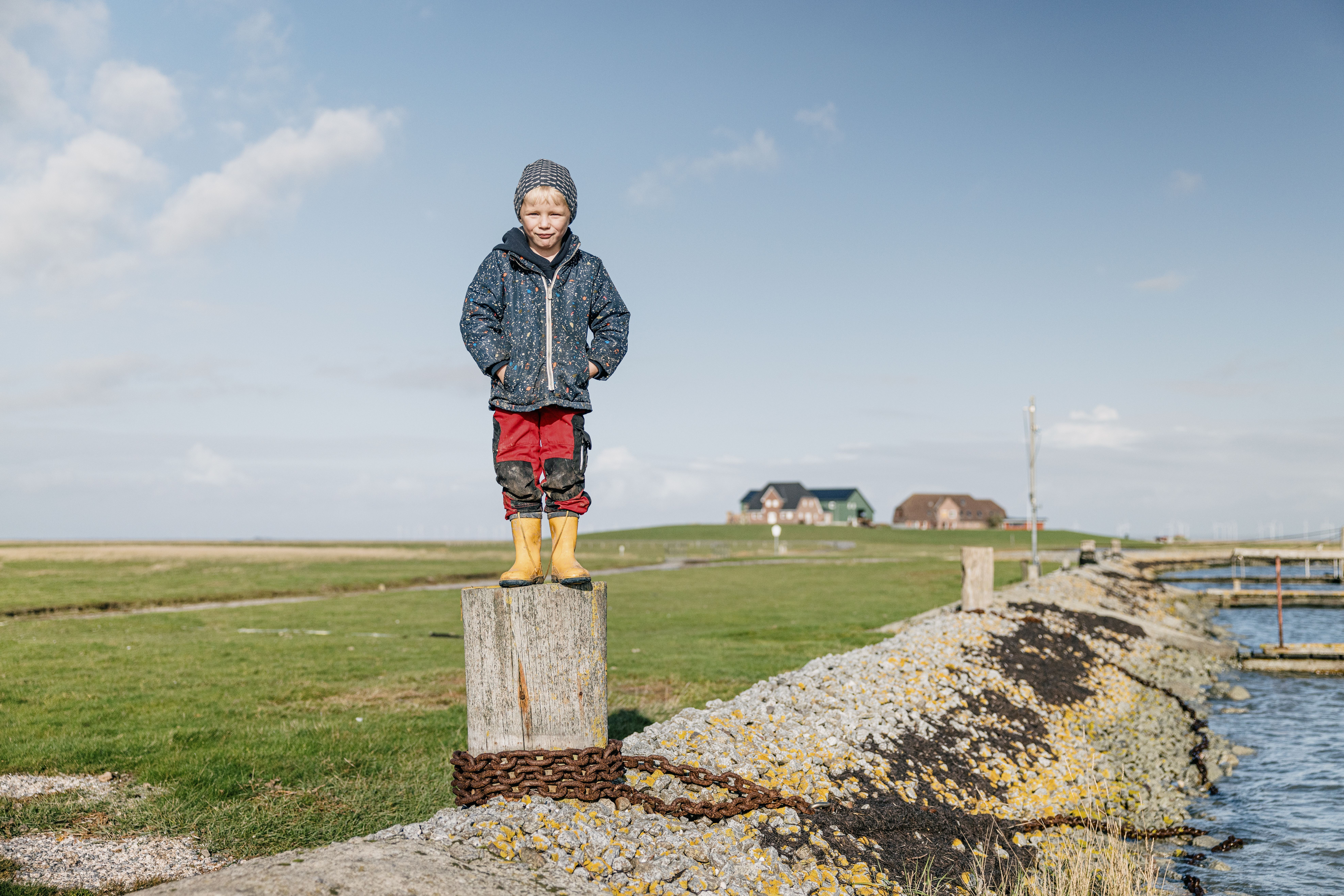 Kind steht auf Pfahl mit gelben Gummistiefeln auf der Hallig Nordstrandischmoor. Verlinkung auf Unterseite "Familienurlaub". 