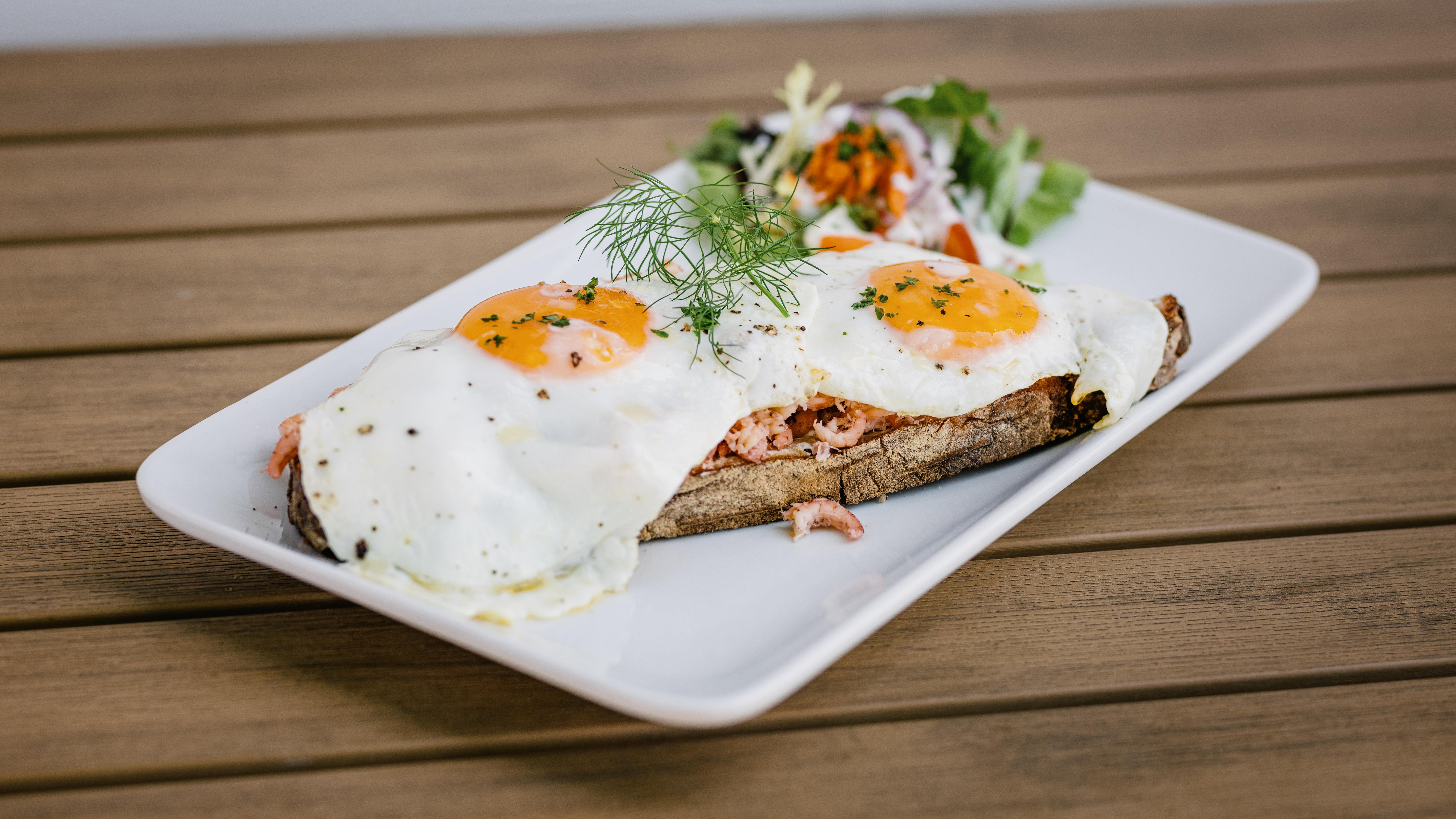 Auf einem viereckigen Teller liegt ein Brot mit Krabben und Spiegelei mit einem kleine Salat auf einem Holztisch. 