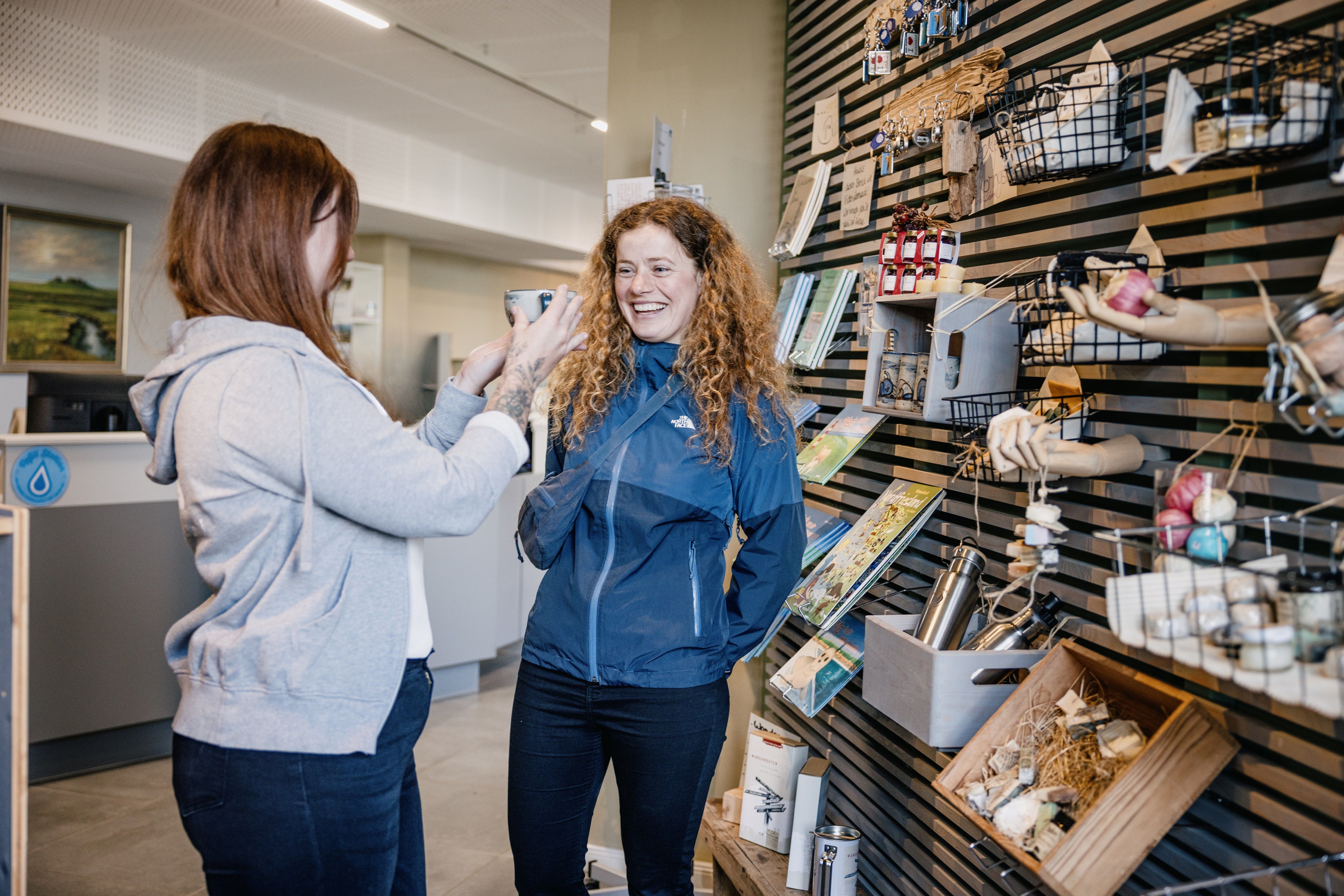 Eine Dame im grauen Pullover zeigt einer weiteren Dame in blauer Jacke eine Tasse.