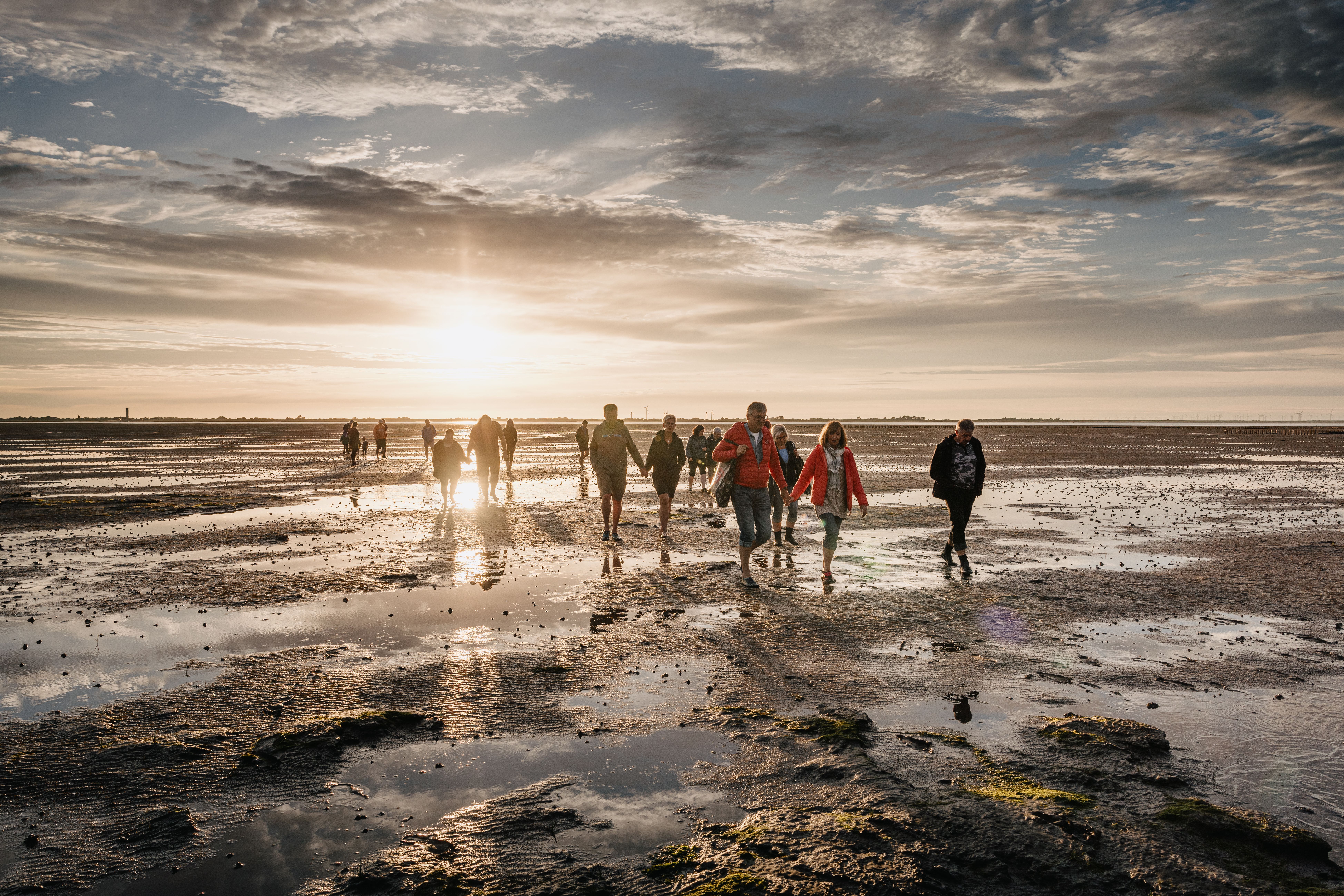 Eine Gruppe läuft durch das schlickige Watt im Sonnenuntergang. Verlinkung zur Unterseite zum Thema "Wattwanderungen auf Nordstrand". 