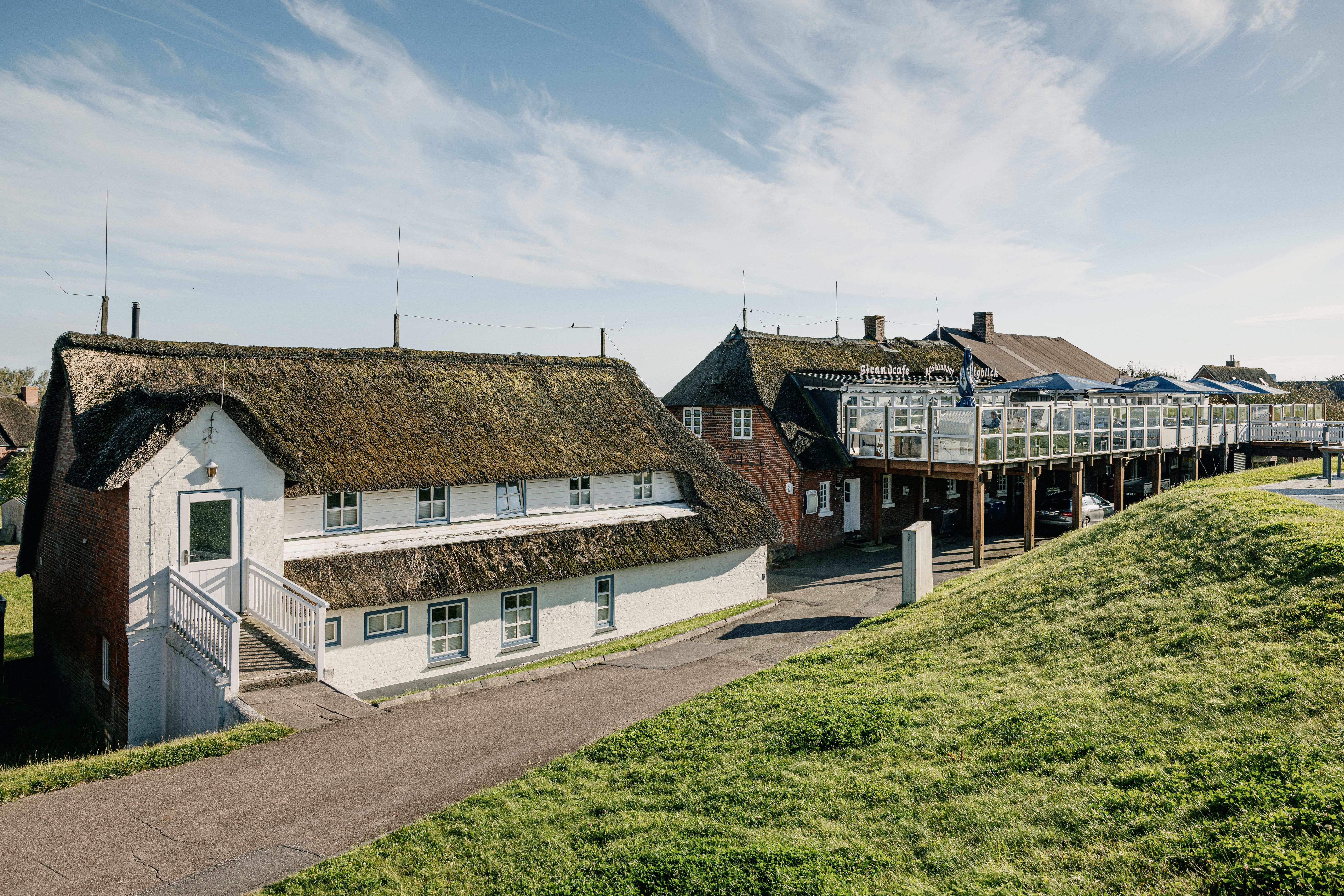 Zwei reetgedeckte Häuser, eins mit weißer Fassade, eins mit Backstein, befinden sich direkt am Deich. Verlinkung auf Unterseite "Hotels und Pensionen auf Nordstrand"