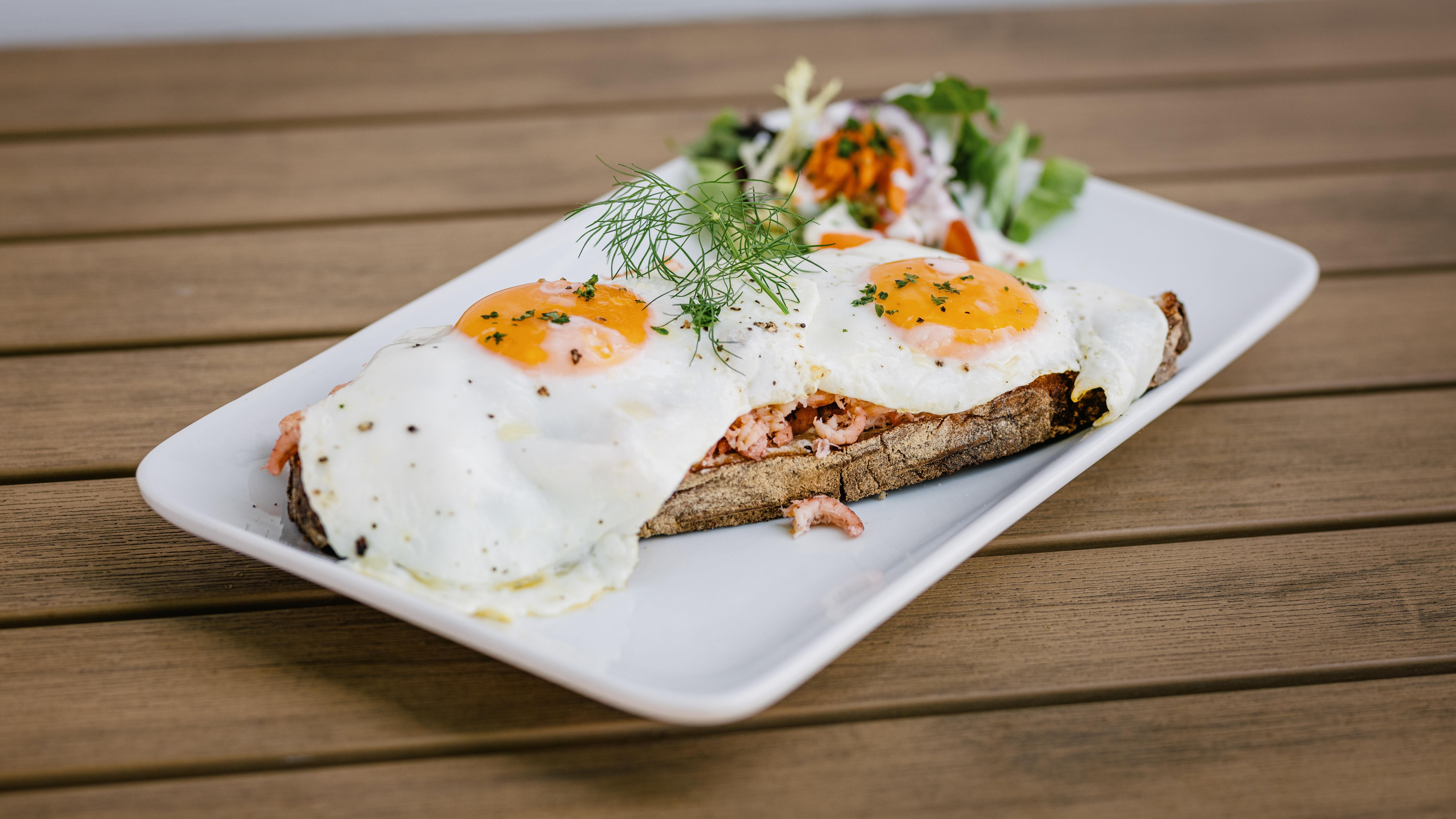 Ein rechteckiger Teller, auf dem sich ein Brot mit zwei Spiegeleiern auf Krabben befindet, ebenso ein kleiner Salat am Tellerrand. Verlinkung auf Unterseite "Gastronomie"