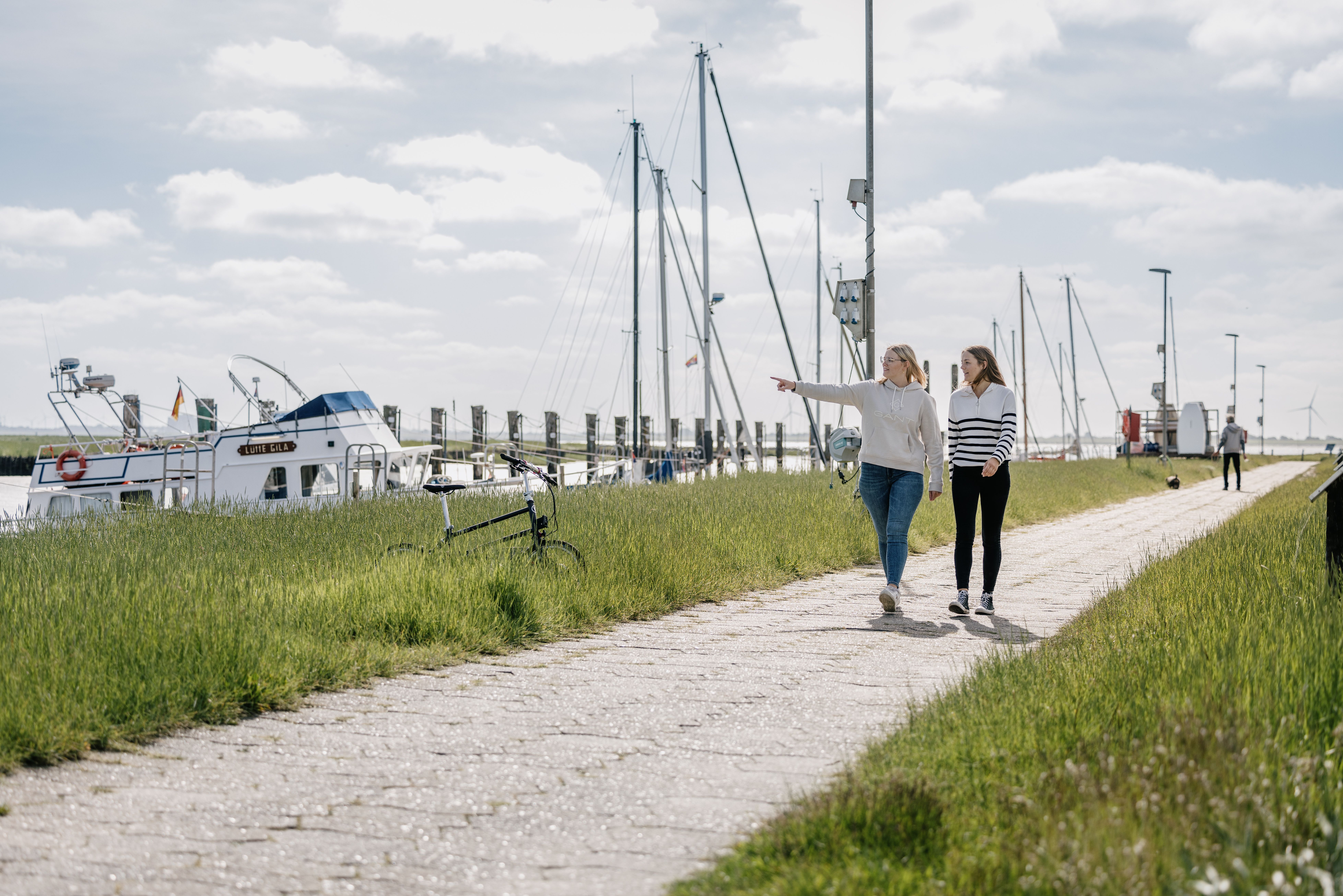 Zwei Frauen spazieren auf einem Weg entlang eines Seglerhafens bei Sonnenschein. Verlinkung auf Unterseite "Ausflugsziele". 